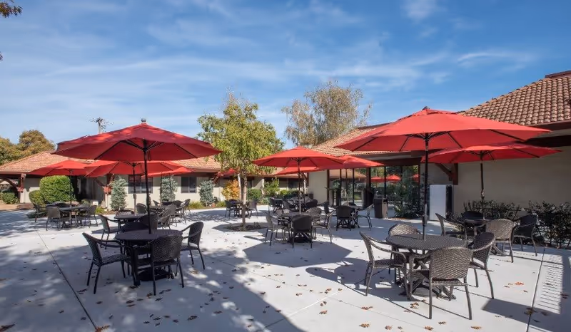 Outdoor patio area at Roseville Commons Senior Living with multiple round tables and chairs shaded by large red umbrellas, surrounded by single-story buildings with tiled roofs and trees under a clear blue sky.