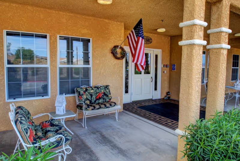 Covered outdoor patio area with beige stucco walls and columns, featuring two cushioned metal benches with floral patterned cushions, a small white table with a decorative lantern, an American flag mounted near a white double door entrance, and some green plants in the foreground.