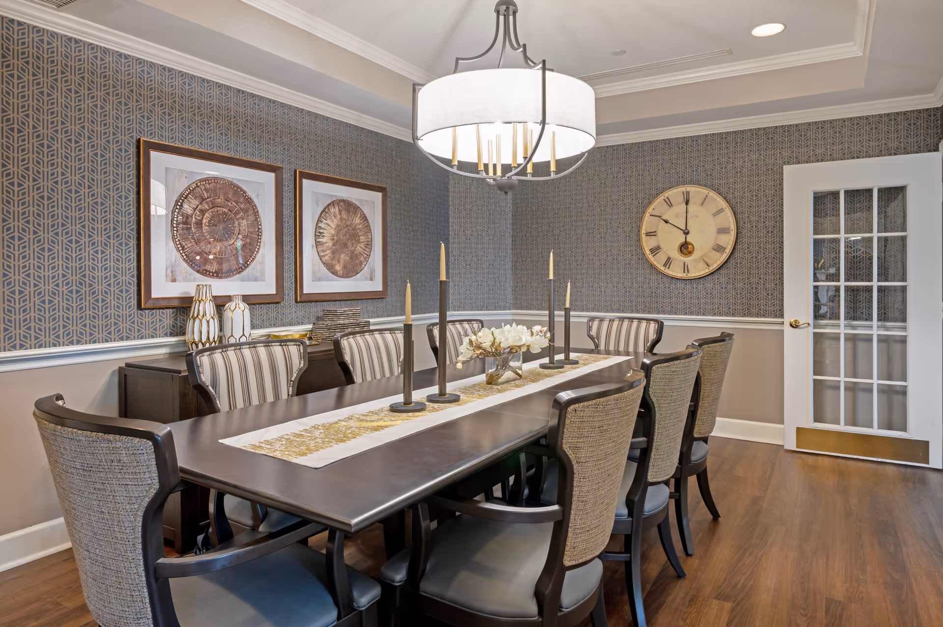 A formal dining room with a long dark wooden table surrounded by eight upholstered chairs. The table is decorated with a white and gold runner, five tall candles, and a small floral arrangement. The walls have a patterned wallpaper with two framed circular art pieces on one side and a large round clock on the adjacent wall. A white door with glass panes is visible in the background, and a modern chandelier hangs above the table.