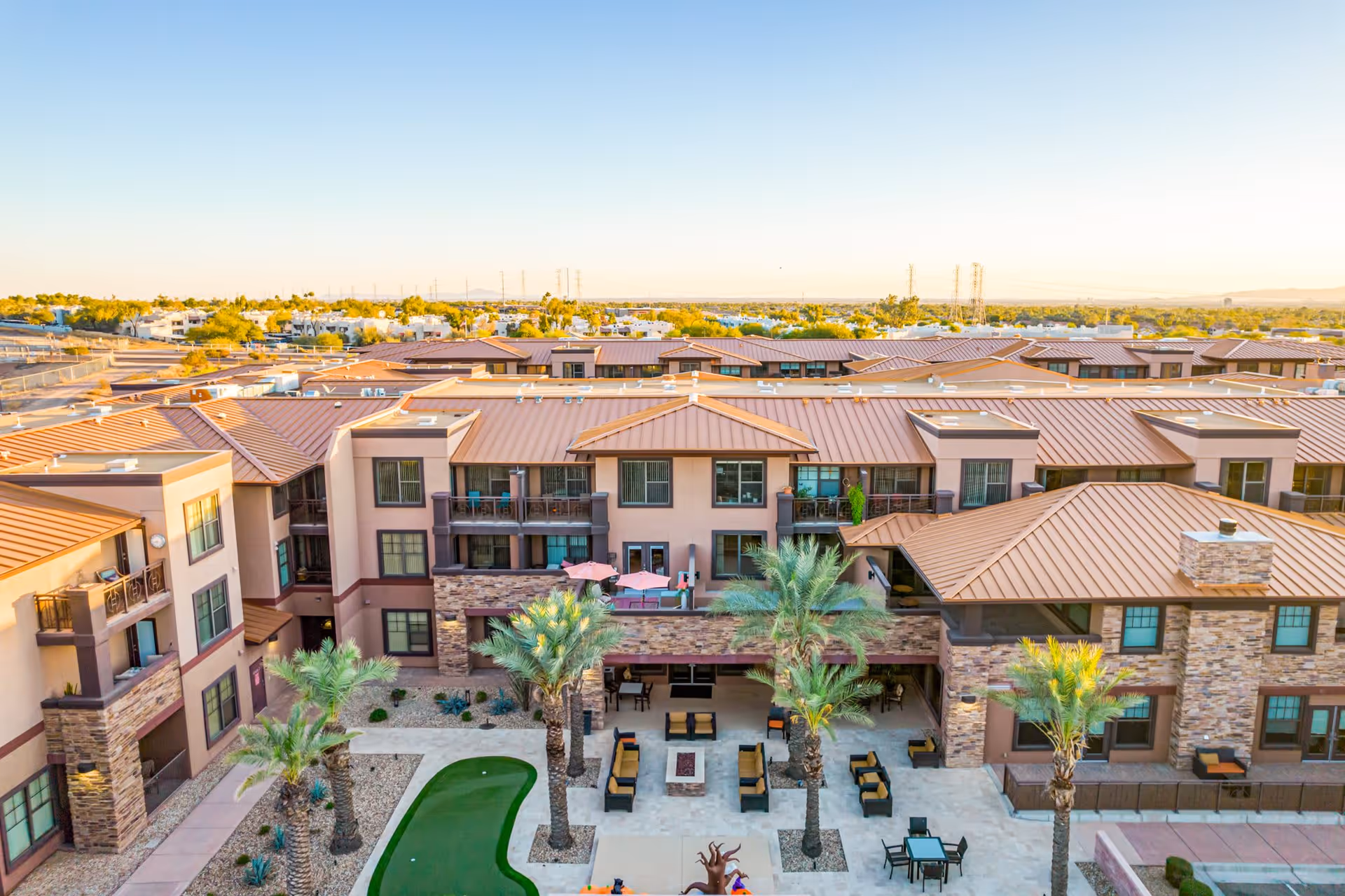 Aerial view of a senior living facility with multiple buildings featuring tan and brown exteriors and metal roofs. The courtyard includes palm trees, outdoor seating areas with chairs and tables, a small artificial putting green, and a fire pit. The surrounding area shows a suburban landscape under a clear sky.