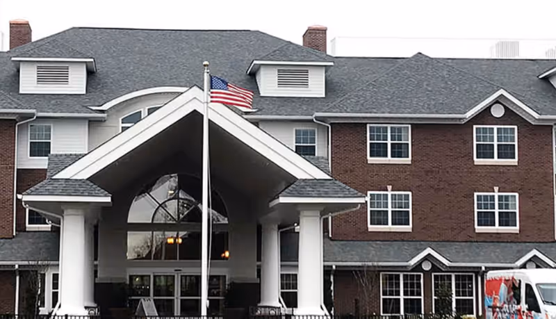 Front exterior view of a multi-story brick building with white trim and a large covered entrance supported by white columns. An American flag is flying on a flagpole in front of the entrance. There is a white van parked on the right side of the building.