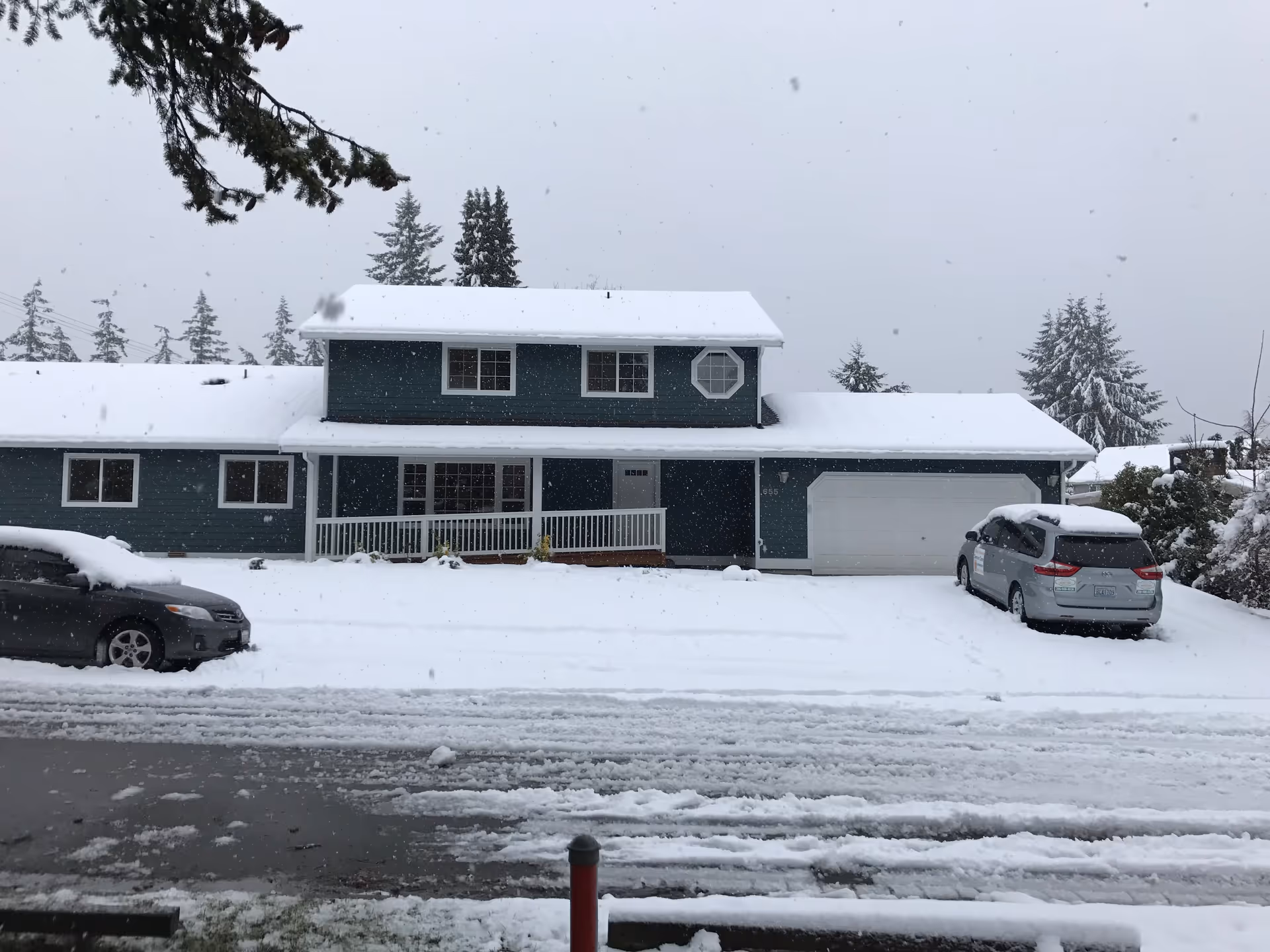 A two-story blue house with a white roof covered in snow during a snowfall. There are two cars parked in the driveway, also covered with snow. Snow-covered trees are visible in the background under a gray sky.
