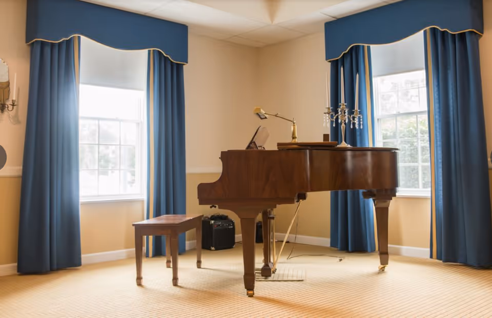 A grand piano and bench sit between two blue-curtained windows in a softly lit room.