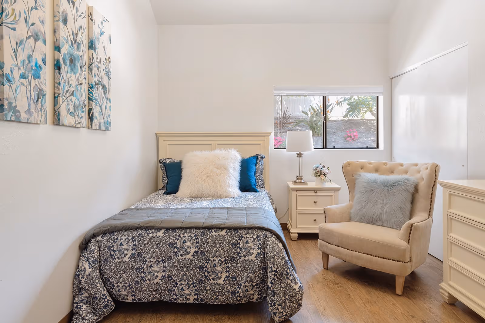 A cozy bedroom with a single bed featuring a blue and white patterned comforter, two blue pillows, and a large white fluffy pillow. Next to the bed is a white nightstand with a lamp and a small flower arrangement. A beige upholstered armchair with a light blue fluffy pillow is positioned near a window showing some greenery and flowers outside. The room has wooden flooring and white walls, with three blue floral paintings hanging on the left wall.
