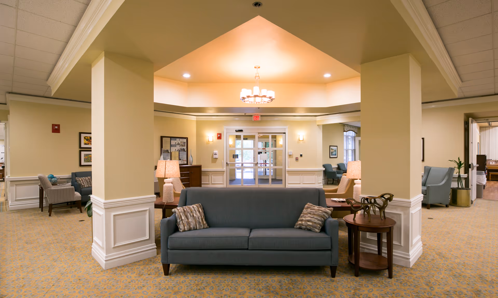 A spacious and well-lit common area in a senior living facility with a blue sofa and patterned cushions in the center, flanked by side tables with lamps. The room features beige walls, carpeted flooring with a patterned design, and decorative columns. In the background, there are additional seating areas with armchairs and framed artwork on the walls, and a set of glass double doors leading outside.