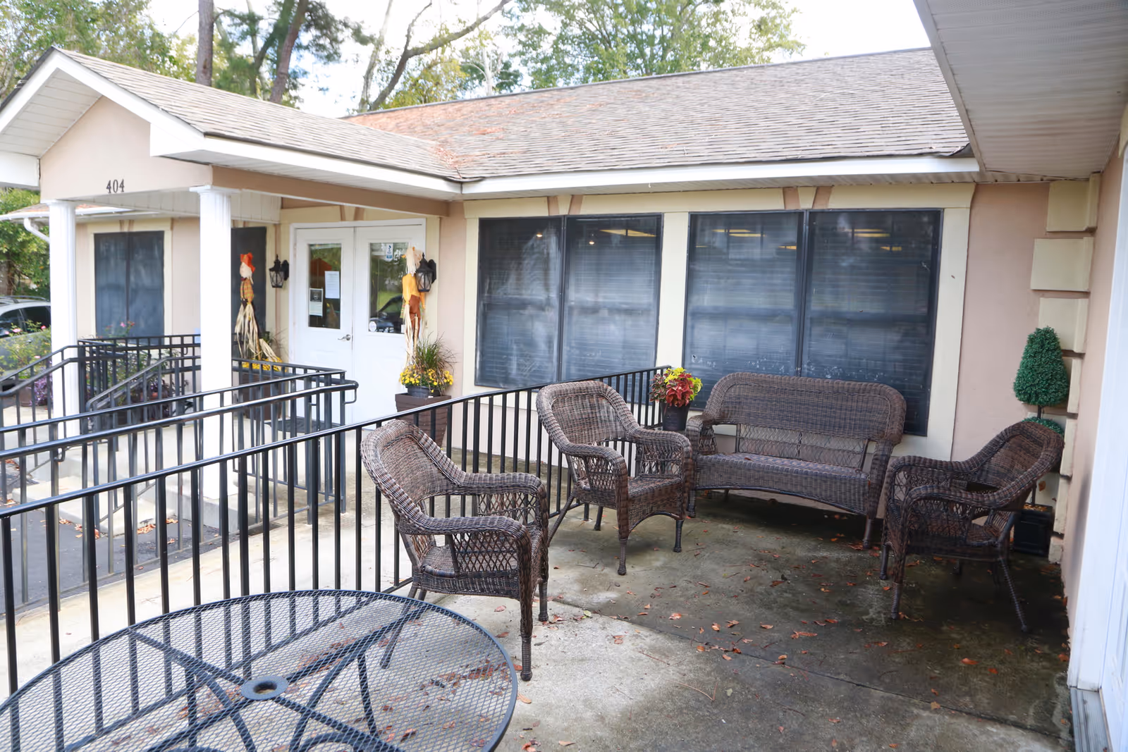 Outdoor patio area at Willow Creek Assisted Living with wicker chairs and a loveseat arranged around a concrete floor. There is a black metal table in the foreground and a building entrance with white double doors and two scarecrow decorations near the door. The building has beige walls and large windows with blinds.