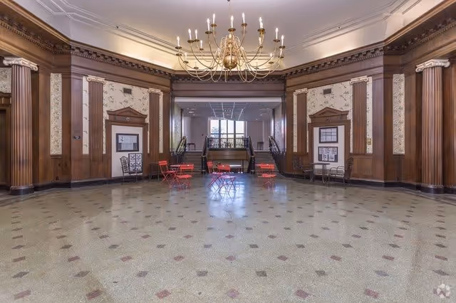 Spacious ornate lobby with wood-paneled columns, a large chandelier, terrazzo floor and red bistro tables and chairs.