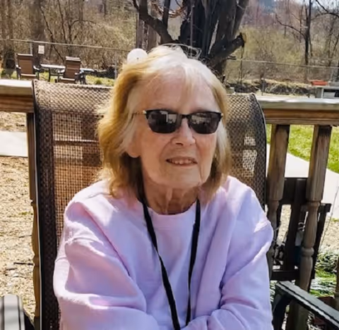 An elderly woman wearing sunglasses and a light purple sweatshirt is sitting outdoors on a mesh chair with a wooden railing and greenery in the background.