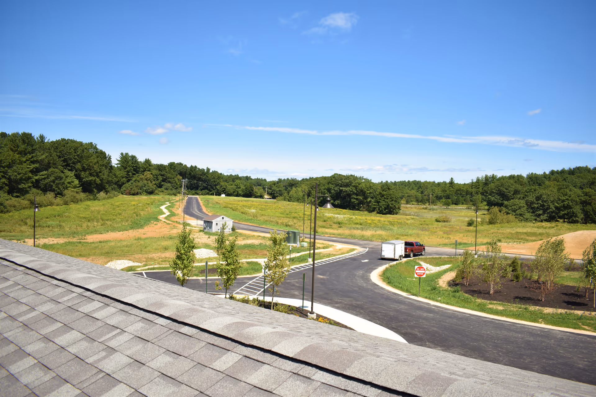 View from a rooftop overlooking a newly paved road with a red truck towing a white trailer, surrounded by green fields, young trees, and a forested area under a clear blue sky.