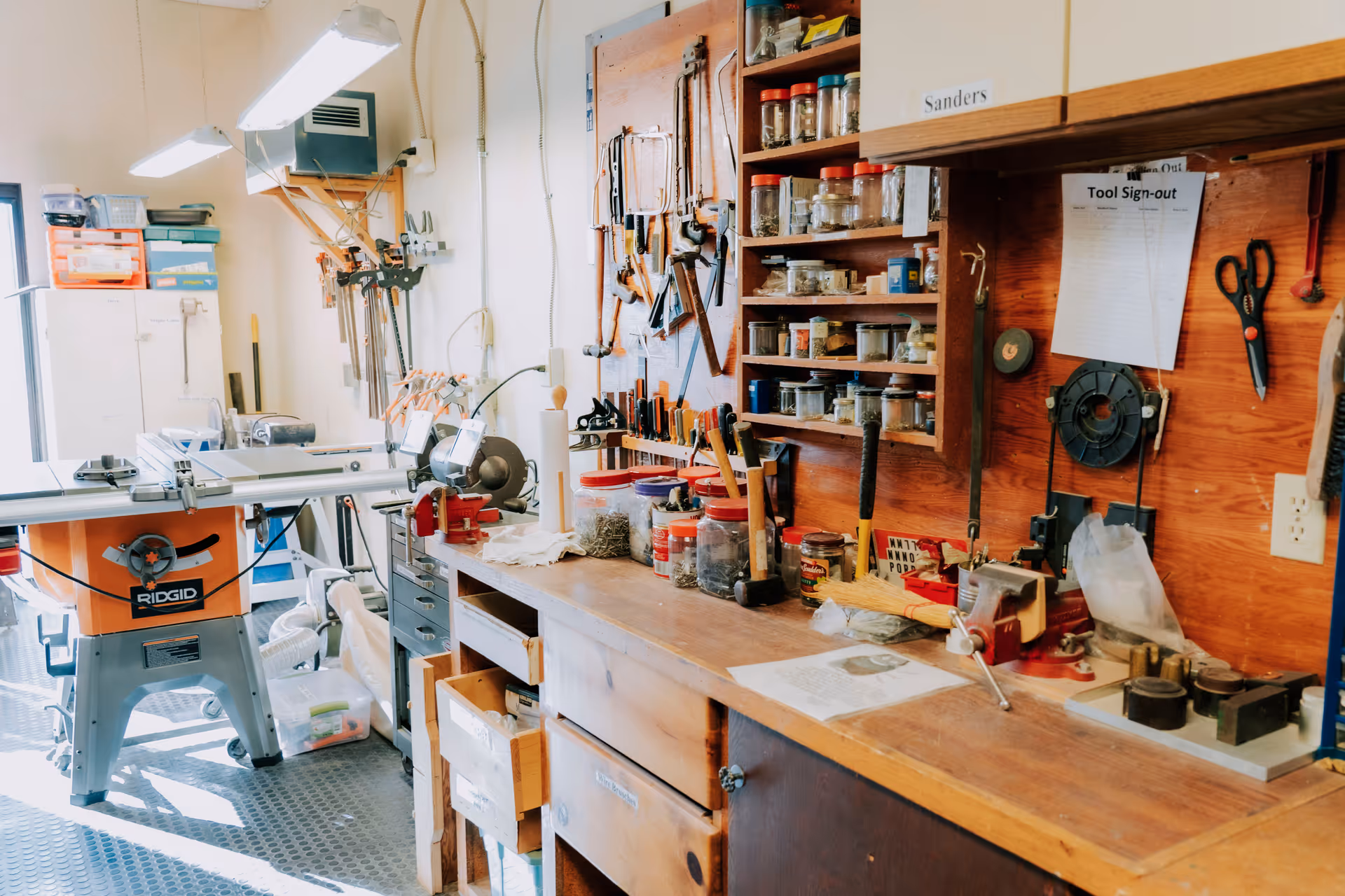 A bright workshop interior with a wooden workbench, pegboard of hand tools and shelves of jars, and a Ridgid table saw.