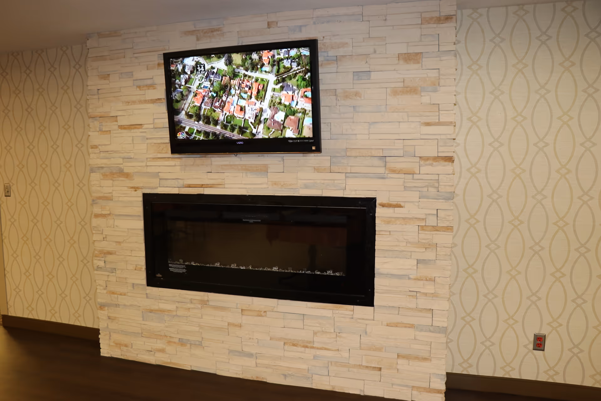 A wall with light-colored stone tiles featuring a mounted flat-screen TV displaying an aerial view of a neighborhood, above a modern black electric fireplace. The surrounding walls have a beige wallpaper with a subtle geometric pattern, and the floor is dark wood.