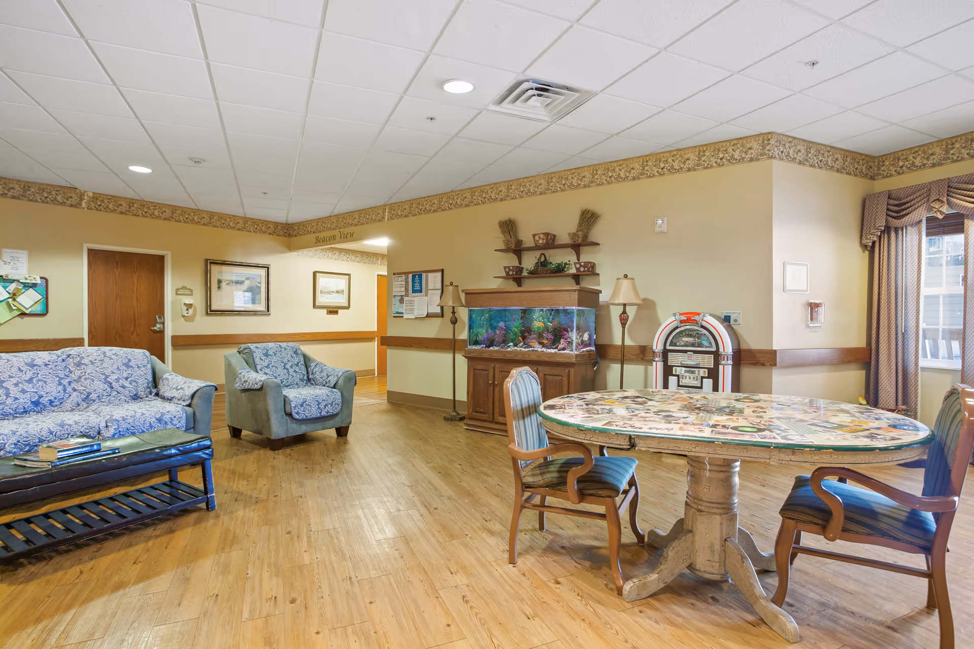 A cozy common area in a senior living facility featuring a wooden floor, a blue patterned sofa and armchair, a round table with two wooden chairs, a large fish tank on a wooden cabinet, a vintage jukebox, and two floor lamps. The walls are beige with a decorative border near the ceiling, and there is a window with drapes letting in natural light.