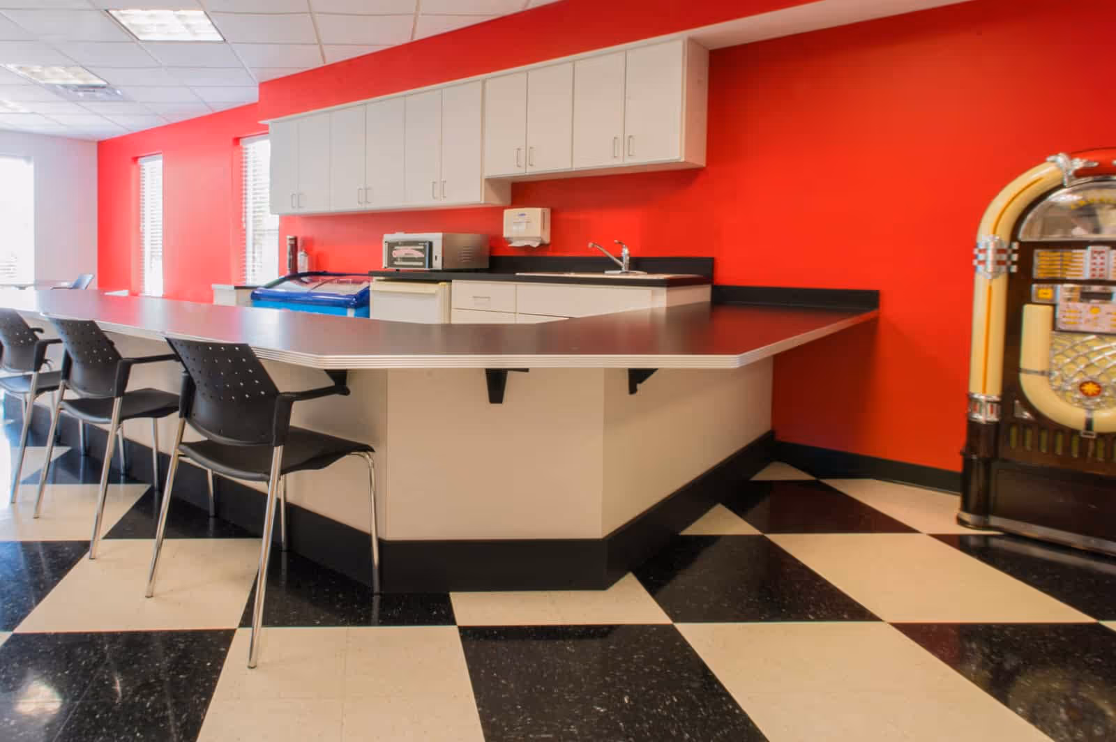 Interior view of a retro-style kitchen or common area with a red wall, white cabinets, a countertop with black chairs, a small sink, a microwave, a freezer, and a vintage jukebox on the right side. The floor has a black and white checkered pattern.