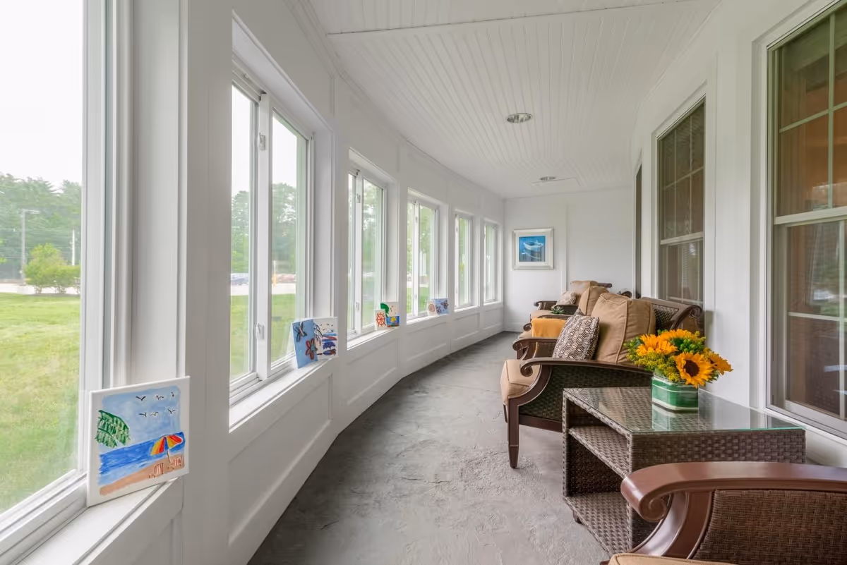 A bright enclosed porch area with large windows along one side, decorated with small paintings on the window sills. The porch has cushioned wicker chairs with patterned pillows and a glass-top wicker side table holding a vase of sunflowers. The walls and ceiling are white, and the floor is a smooth gray surface.
