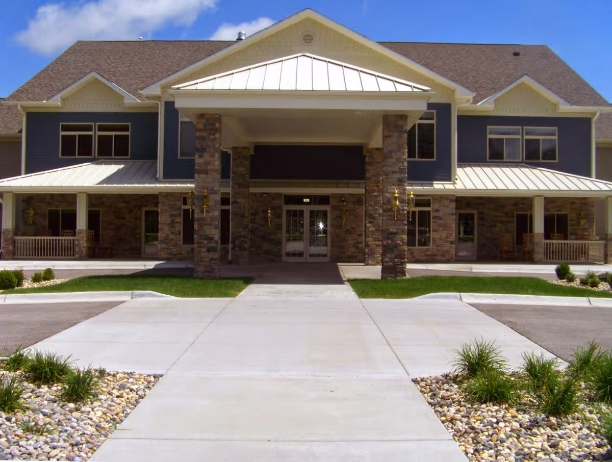 Front entrance of a two-story building with a covered porte-cochere, stone pillars, walkways and landscaped rock beds.