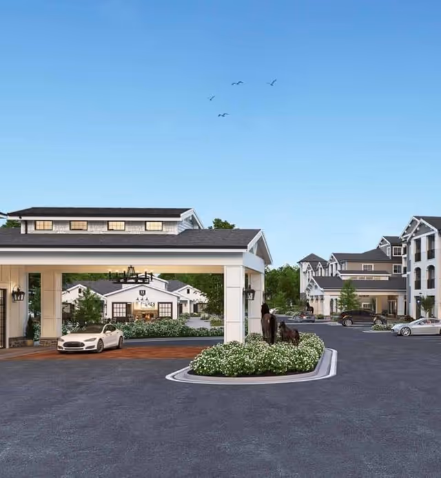 Entrance area of The Park at University Village Independent Living Community featuring a covered drop-off zone with a white car parked underneath, landscaped flower beds, statues of horses, and multi-story residential buildings in the background under a clear blue sky with birds flying.