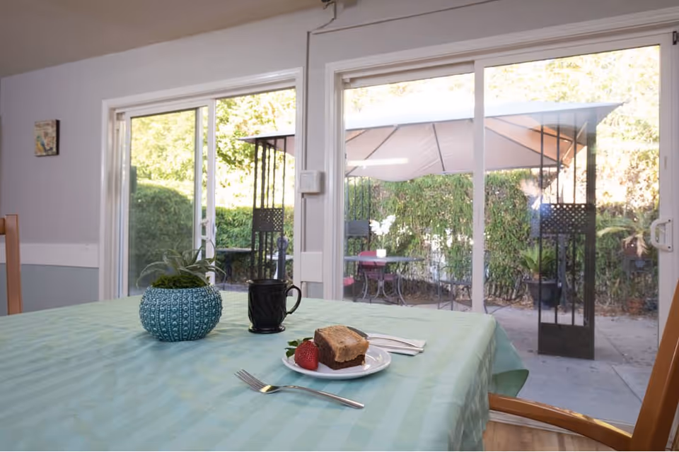 A dining table covered with a light green tablecloth with a small potted plant, a black coffee mug, and a plate with a piece of cake and a strawberry. In the background, large sliding glass doors open to an outdoor patio area with a table, chairs, and a canopy, surrounded by greenery.