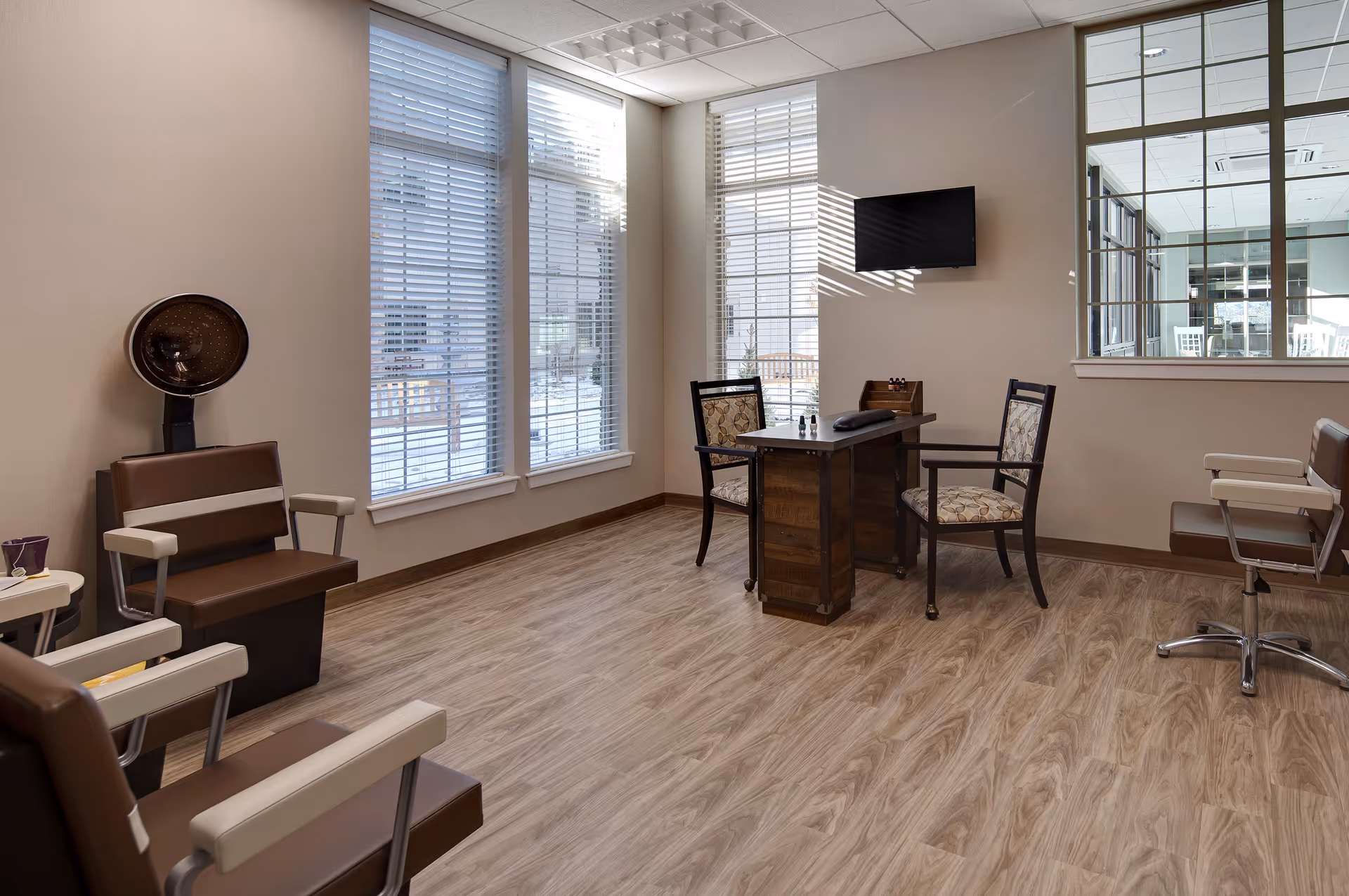 A bright and clean salon room with large windows covered by white blinds letting in natural light. The room features several brown and beige salon chairs, a hair dryer chair, a wooden manicure table with two patterned chairs, and a wall-mounted flat screen TV. The floor has a light wood finish and the walls are painted beige.