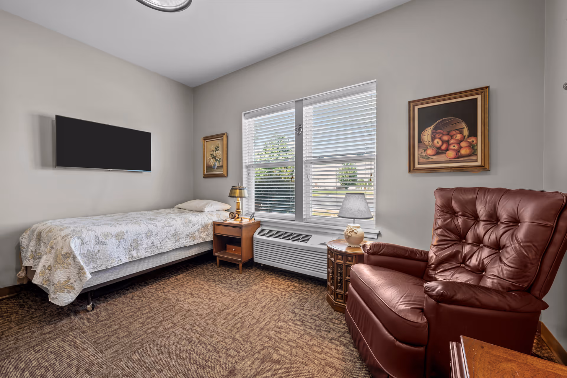 A senior living bedroom with a single bed covered in a floral quilt, a wooden nightstand with a lamp, a large window with blinds, a wall-mounted flat screen TV, two framed paintings on the walls, and a brown leather armchair next to a small round wooden table with a lamp.