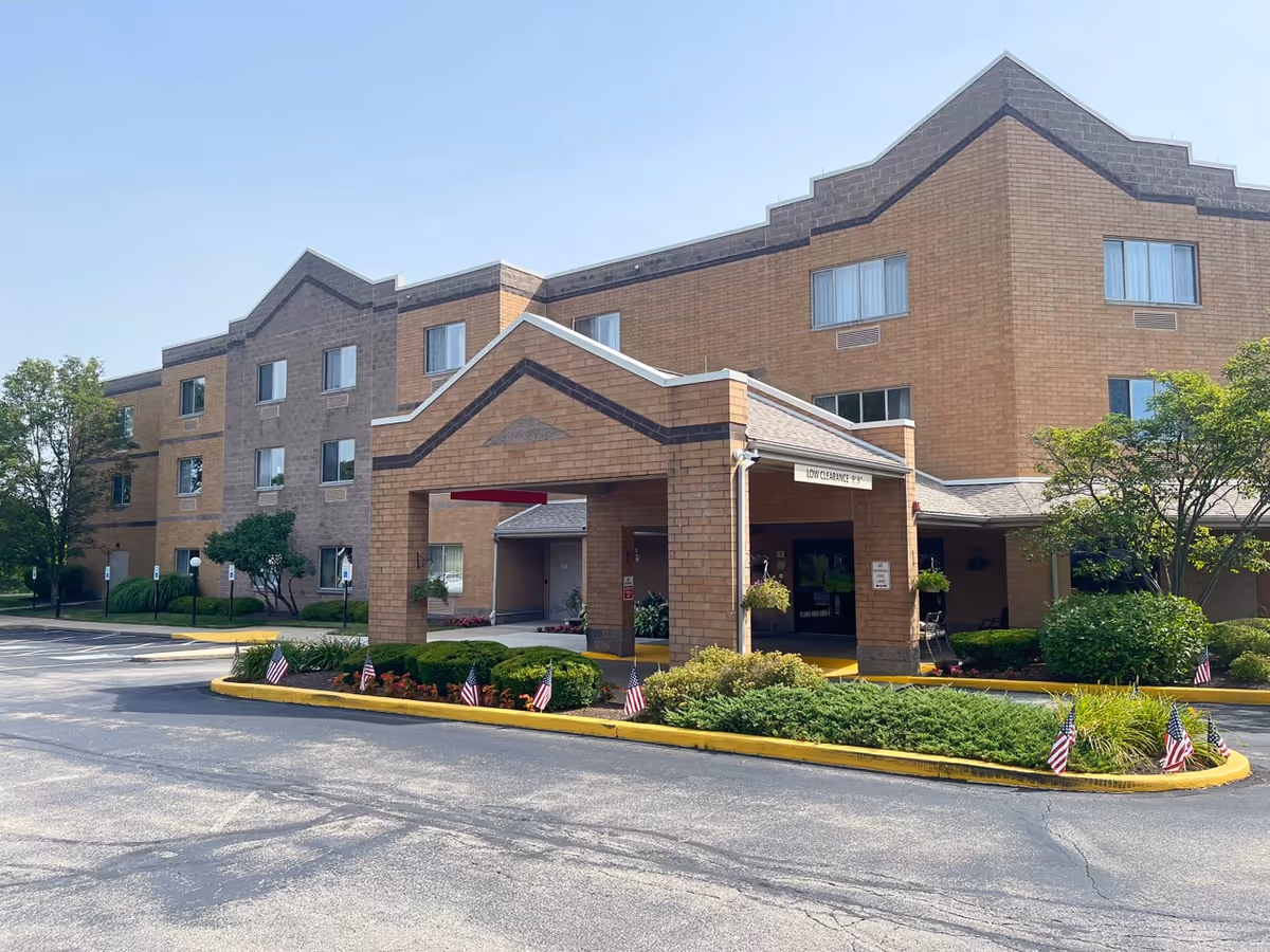 Exterior view of a three-story brick building with a covered entrance and landscaped greenery. Small American flags are placed along the curb in front of the entrance. The sky is clear and blue.