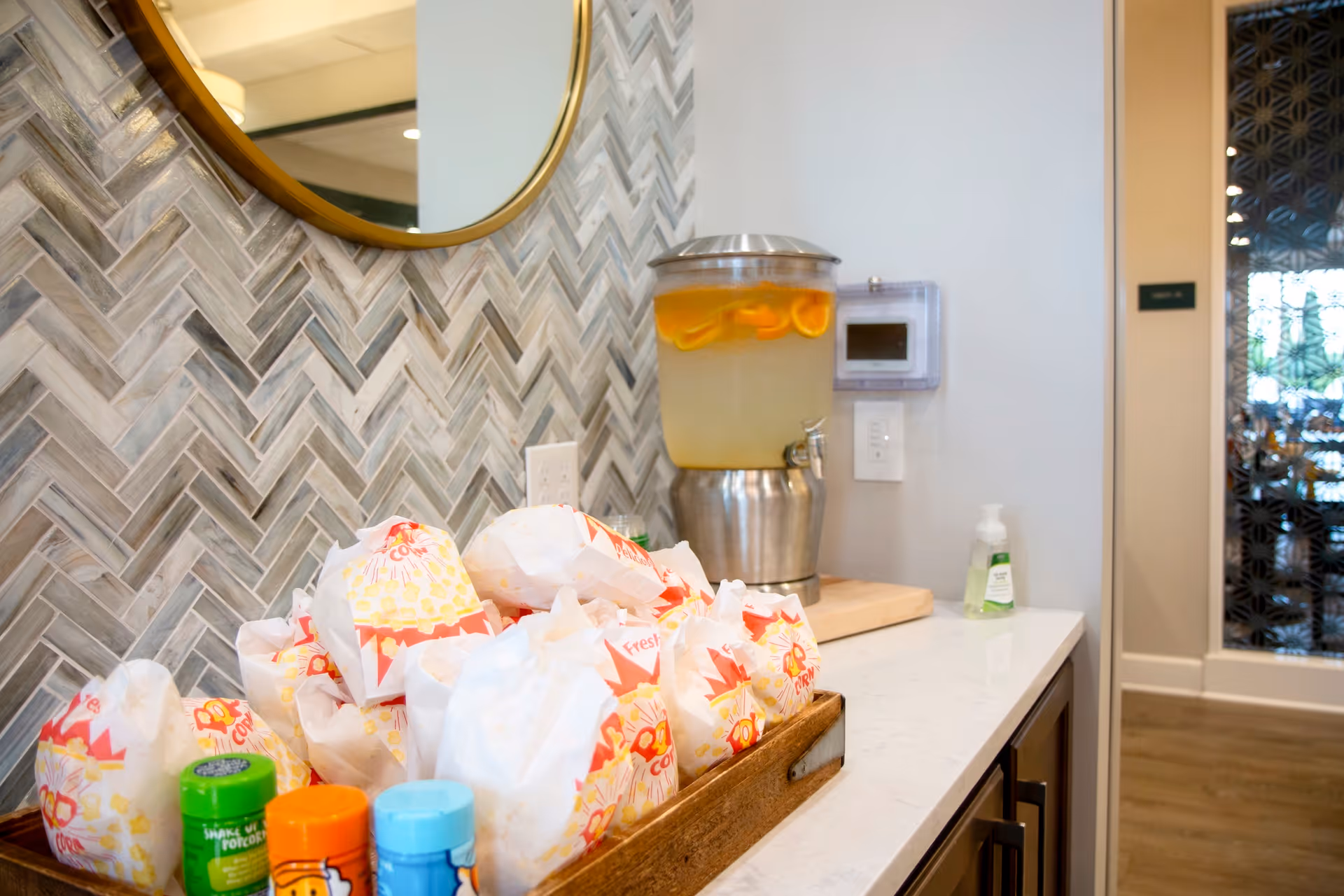 A countertop with a wooden tray filled with popcorn bags, several seasoning containers, a large beverage dispenser filled with lemon water, and a bottle of hand sanitizer. The background features a herringbone patterned tile wall and a round mirror.