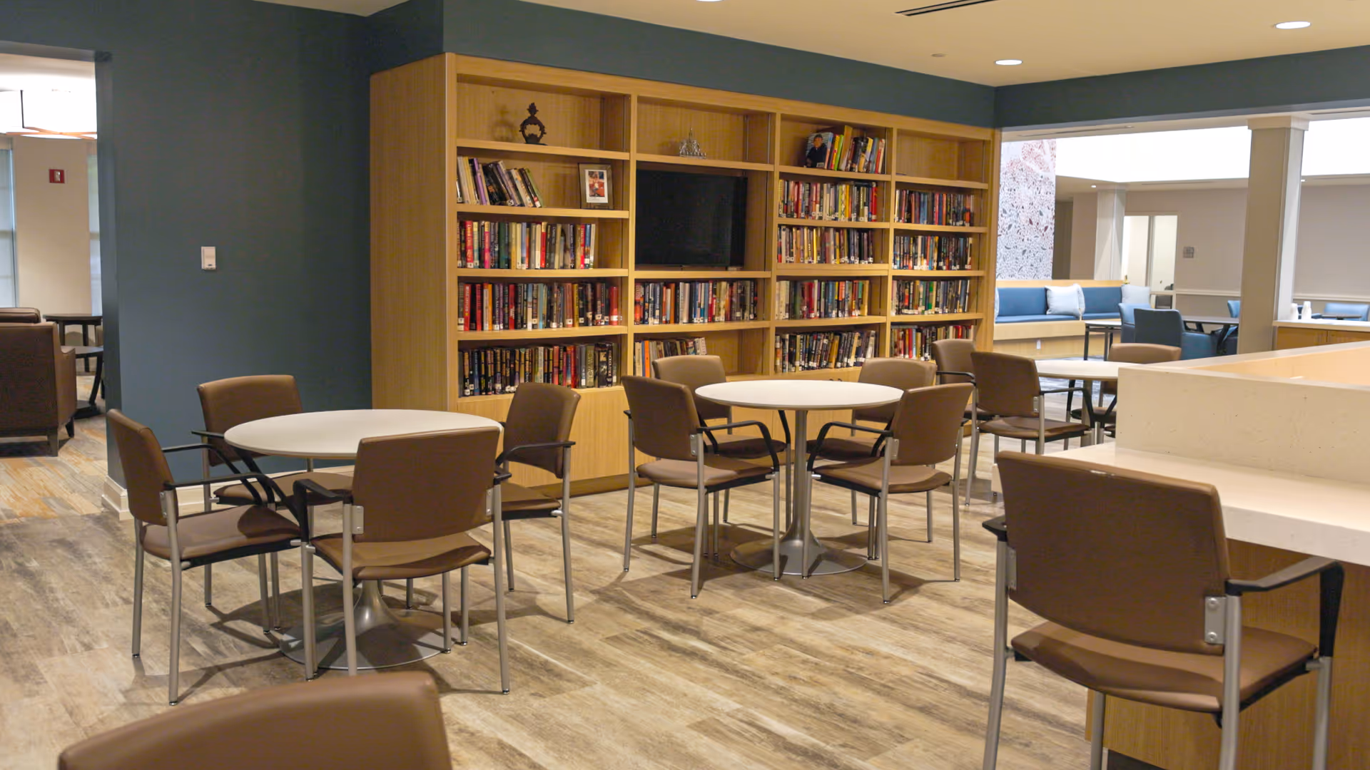 A senior living facility common area with round tables and brown chairs arranged for seating. A large wooden bookshelf filled with books and a TV is mounted on the wall. The room has wood-style flooring and neutral-colored walls, with additional seating visible in the background.