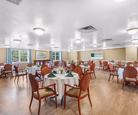 Bright dining room with round tables set with white tablecloths, green napkins, and wooden chairs.