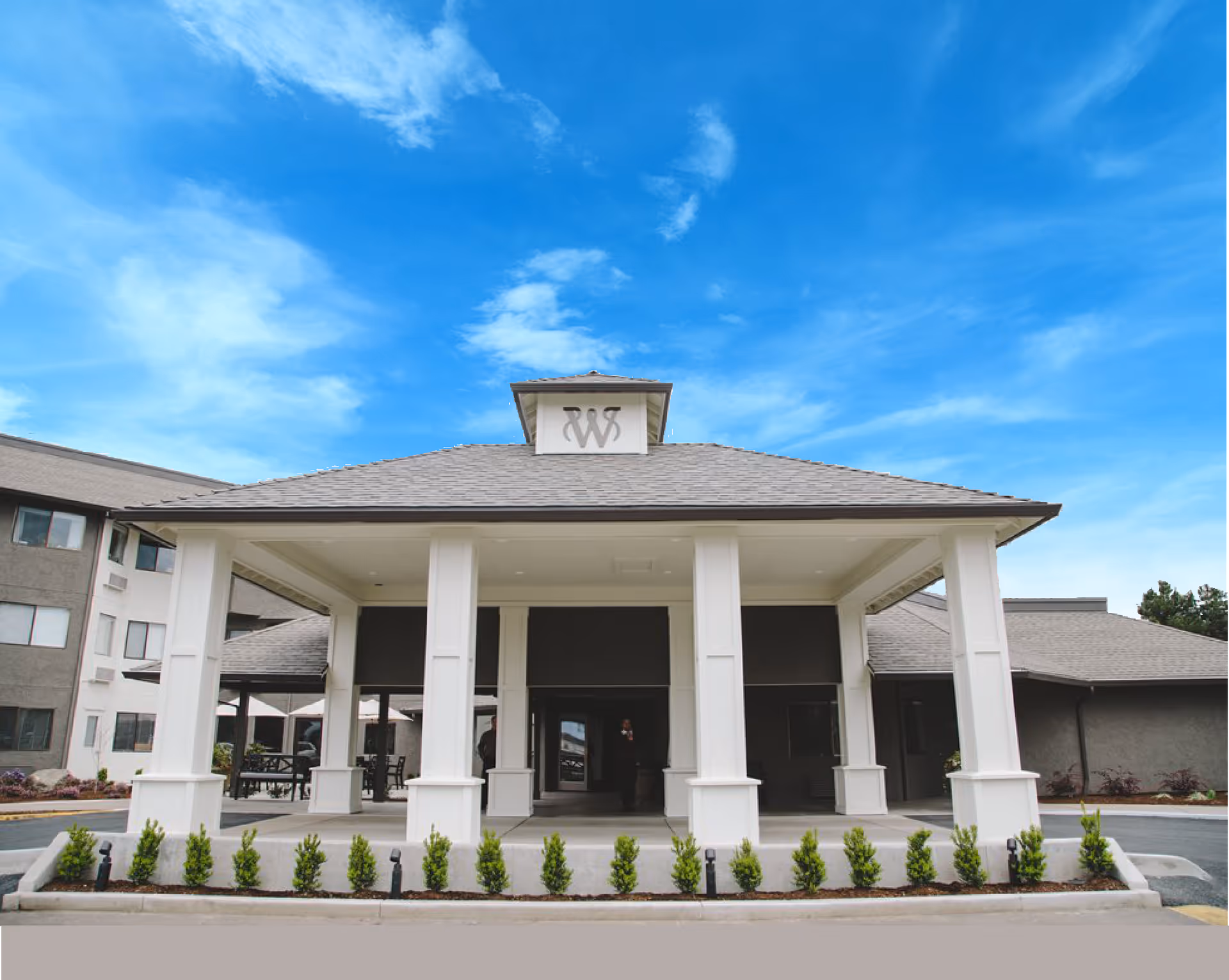 Front exterior view of Weatherly Inn Independent Senior Living building entrance with white pillars, a covered drop-off area, and a clear blue sky above.