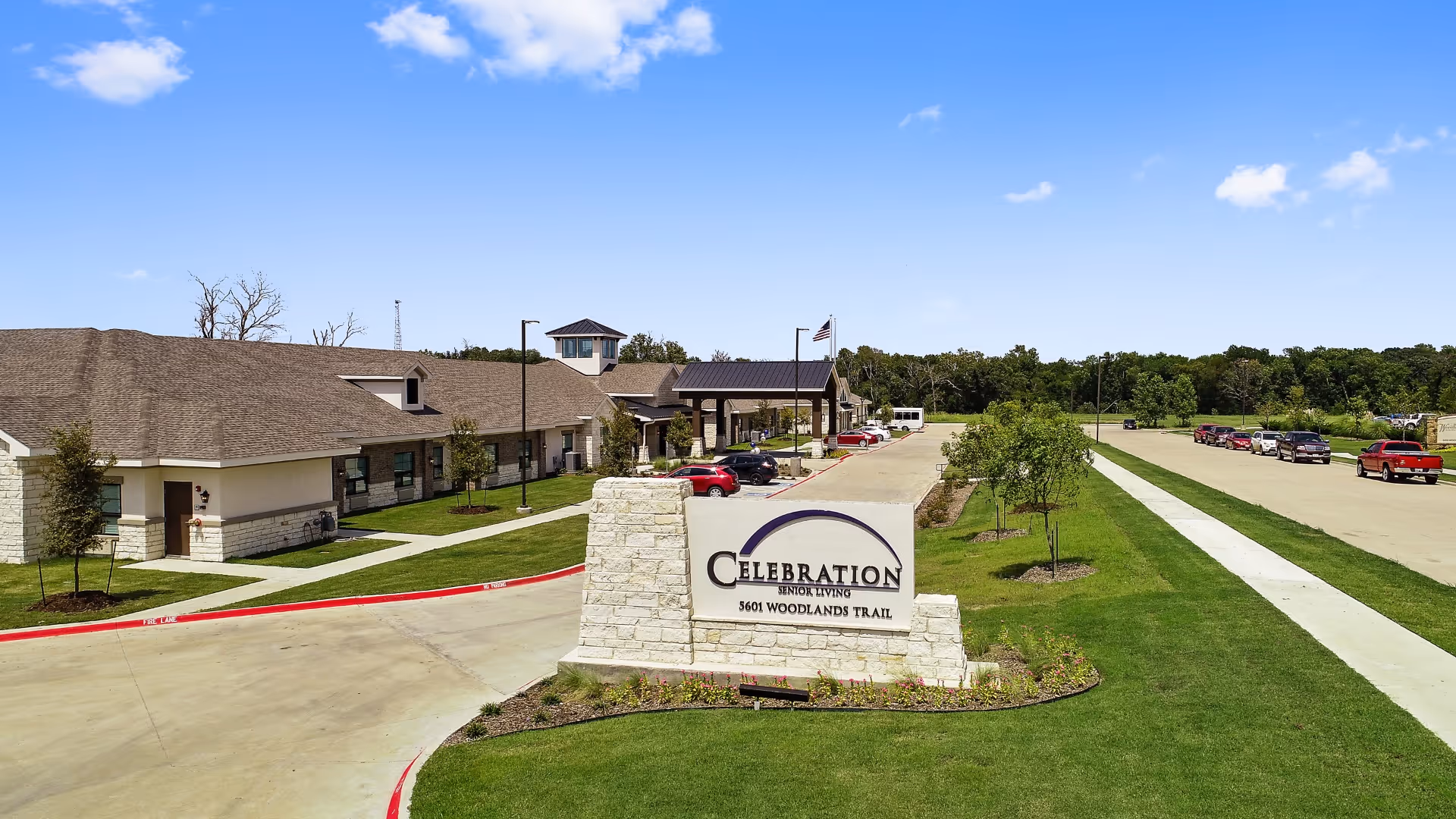 Exterior view of Celebration Senior Living of Denison facility on a sunny day, showing the building, parking lot with several cars, a sidewalk, and a sign with the facility name and address at the entrance.