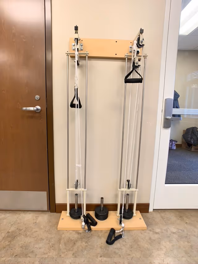 A wall-mounted pulley exercise machine with weights and handles, positioned between a wooden door and a glass door in an indoor room with beige walls and a tiled floor.