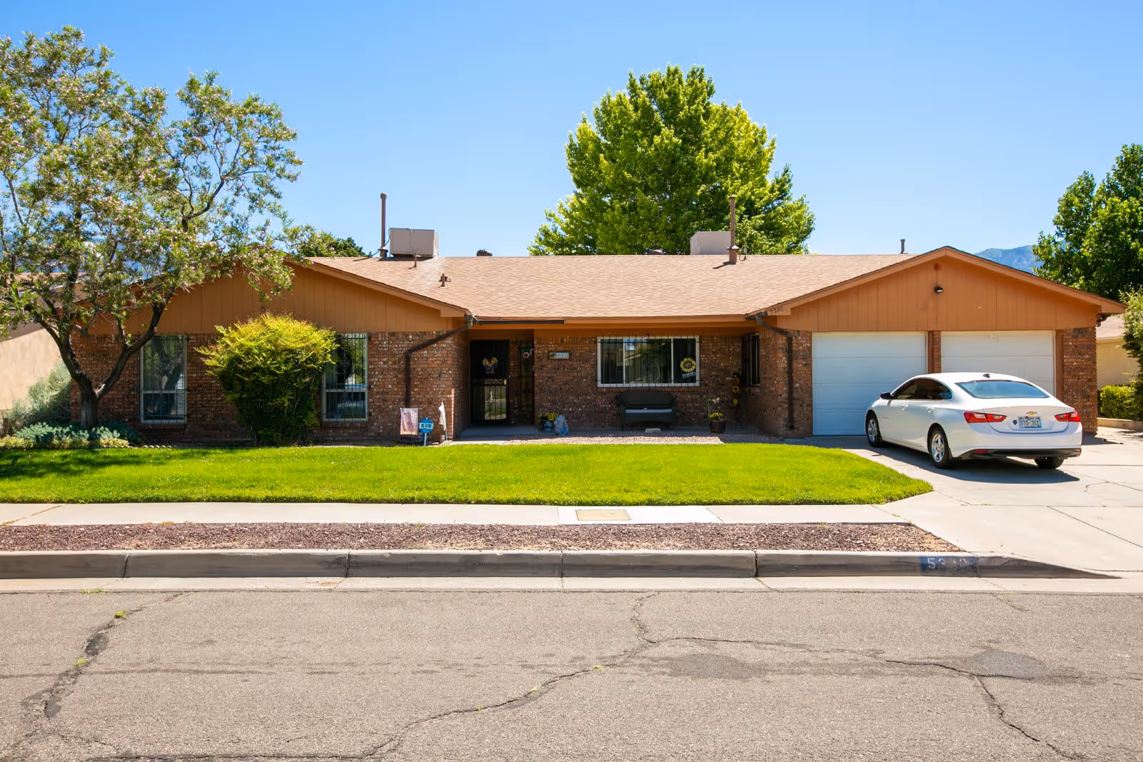 Single-story brick house with a brown roof, green lawn, a tree on the left, and a white car parked in the driveway under a clear blue sky.