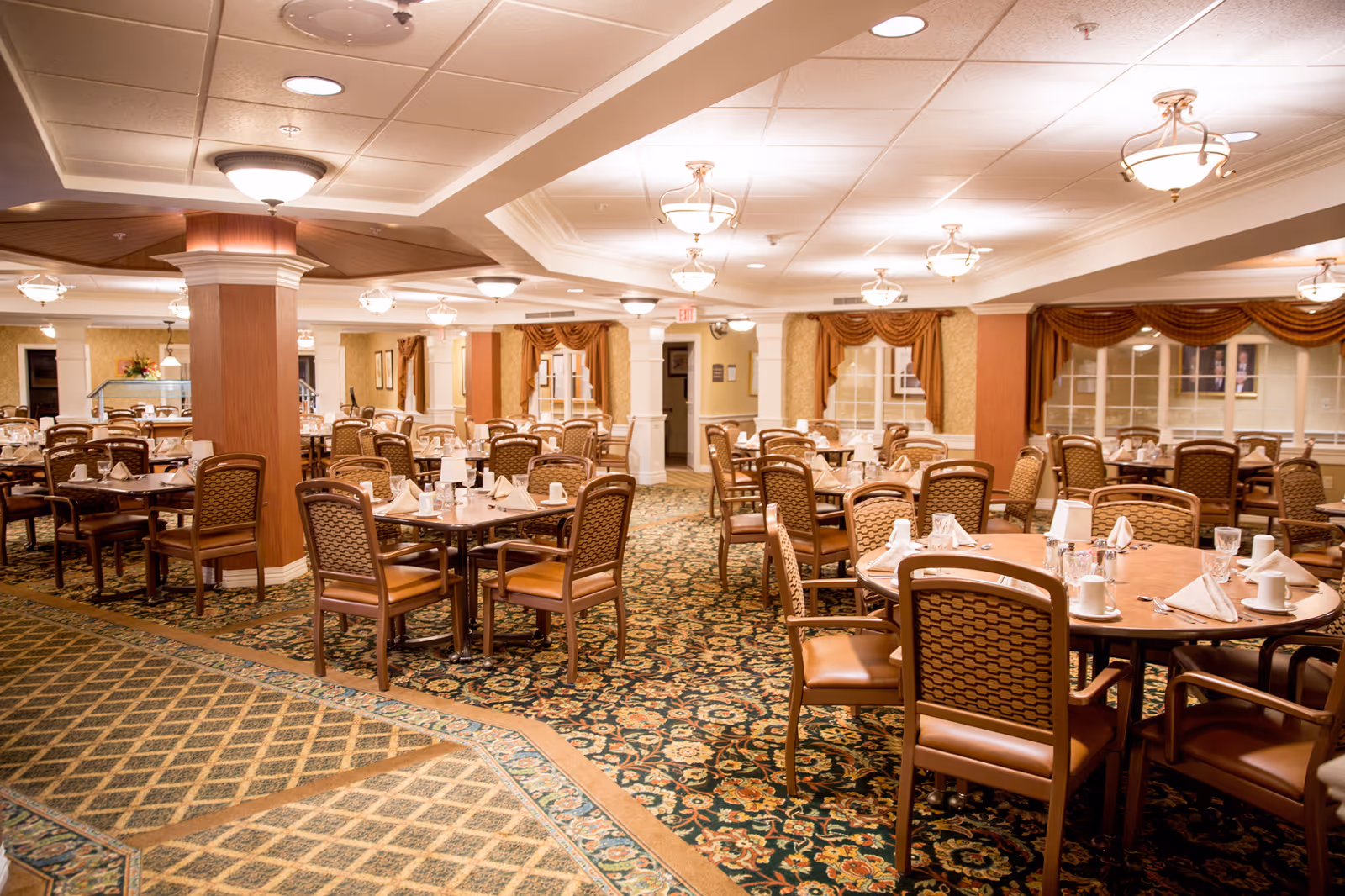 A large, carpeted dining room with multiple round and square tables set with napkins and glassware, surrounded by wooden chairs and warm overhead lighting.