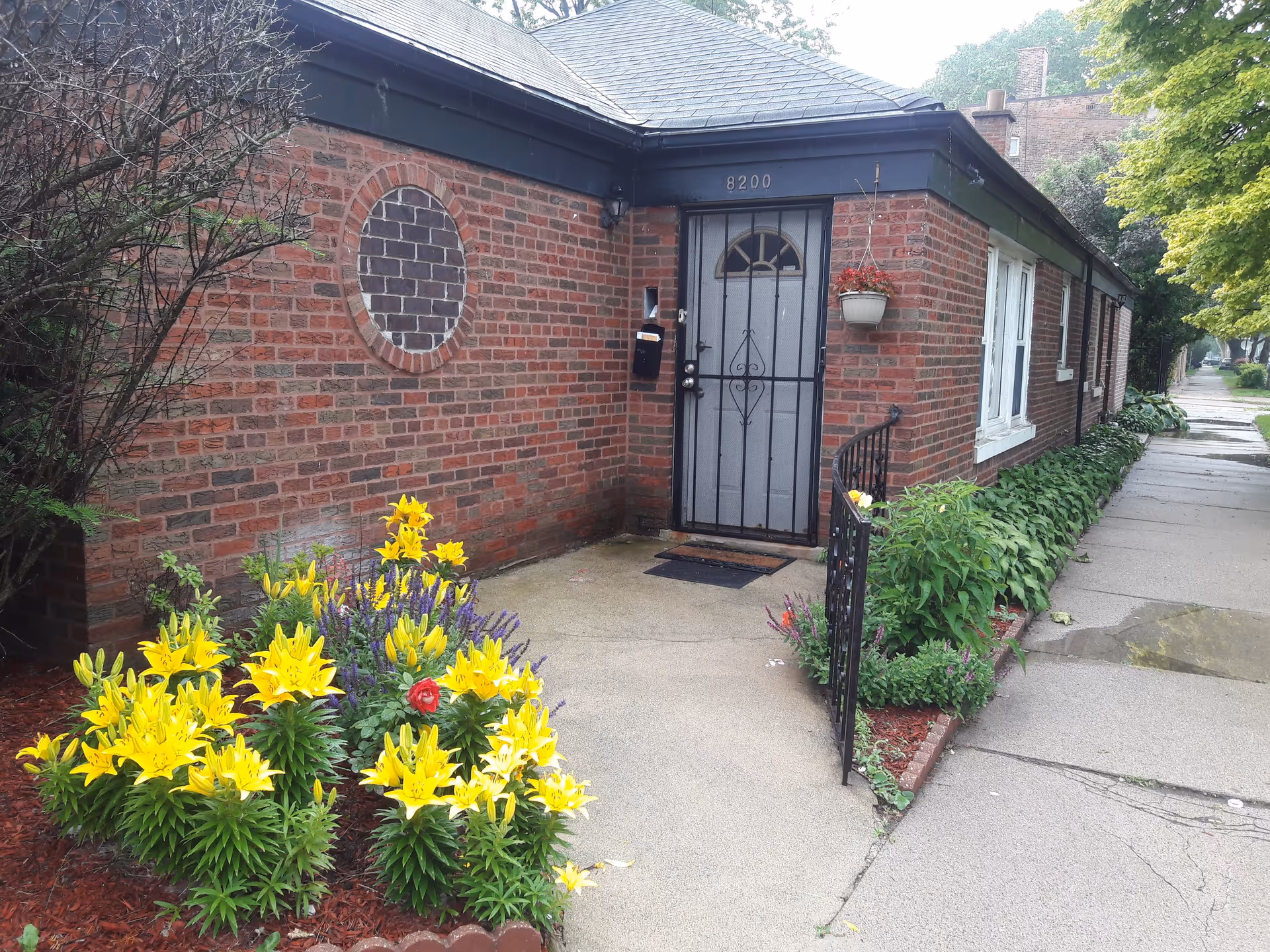 Exterior view of a brick building with the address number 8200 above a black metal door with a screen. There is a small garden bed with yellow flowers and other plants near the entrance, and a sidewalk runs alongside the building. A hanging flower pot with red flowers is mounted on the wall next to the door.