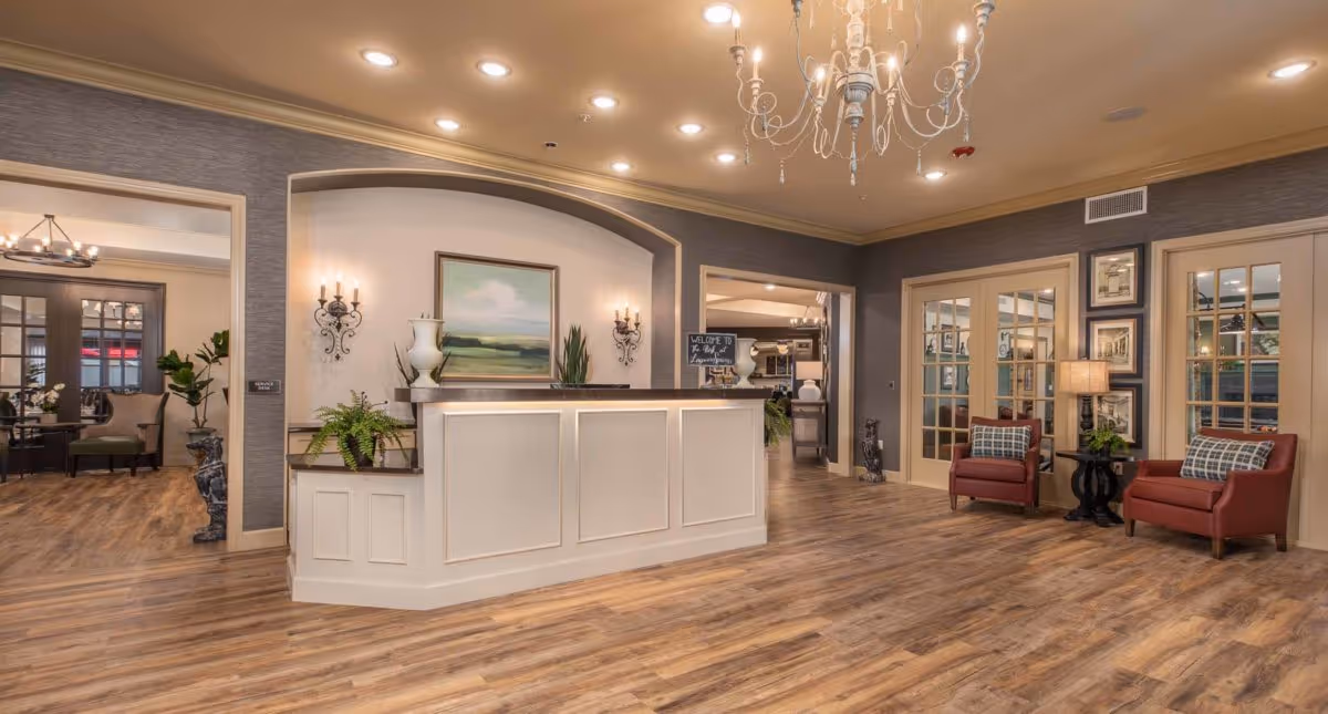 Spacious senior living reception area with a white front desk, chandelier, wood floors, and seating alcoves.