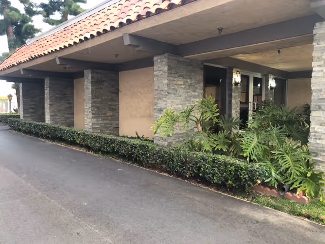 Exterior view of a building with a covered walkway supported by stone pillars, a tiled roof, and landscaped greenery including bushes and large leafy plants along the side.