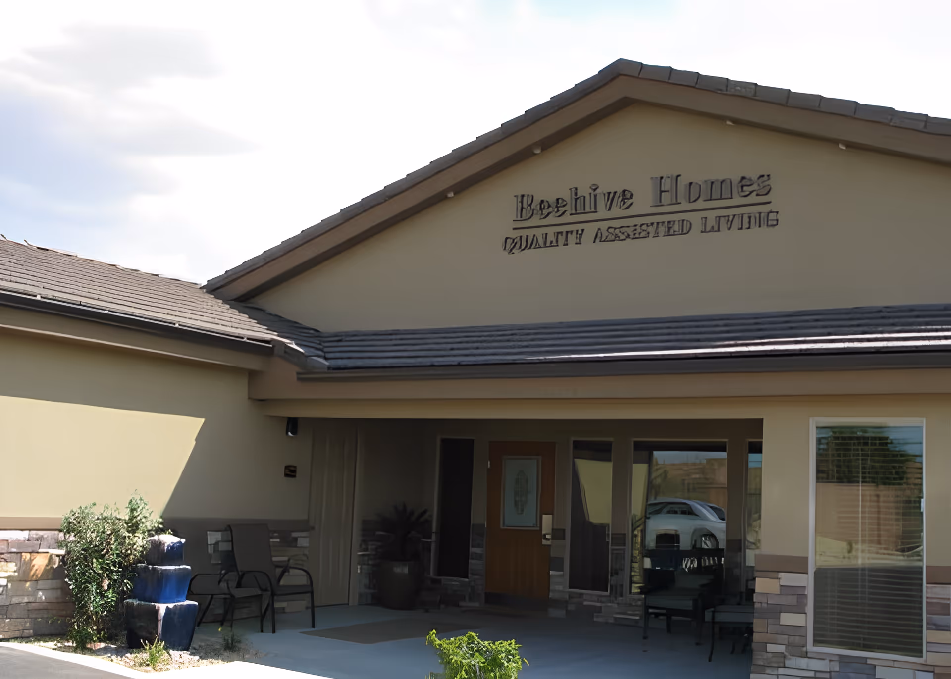 Exterior front view of Beehive Homes Of Washington County 5 building with a covered entrance, outdoor chairs, potted plants, and a sign that reads 'Beehive Homes Quality Assisted Living'.