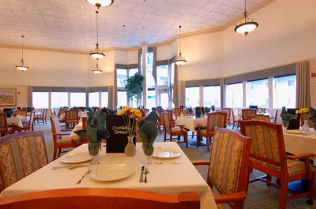 Spacious dining room with neatly set tables and chairs, chandeliers, and large windows in a senior living facility.