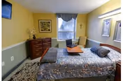 A cozy bedroom with a bed covered in a patterned blue and gray bedspread, a wooden dresser with a vase and flowers, a window with curtains, a cushioned chair, framed artwork on the walls, and a small cross hanging near the window.