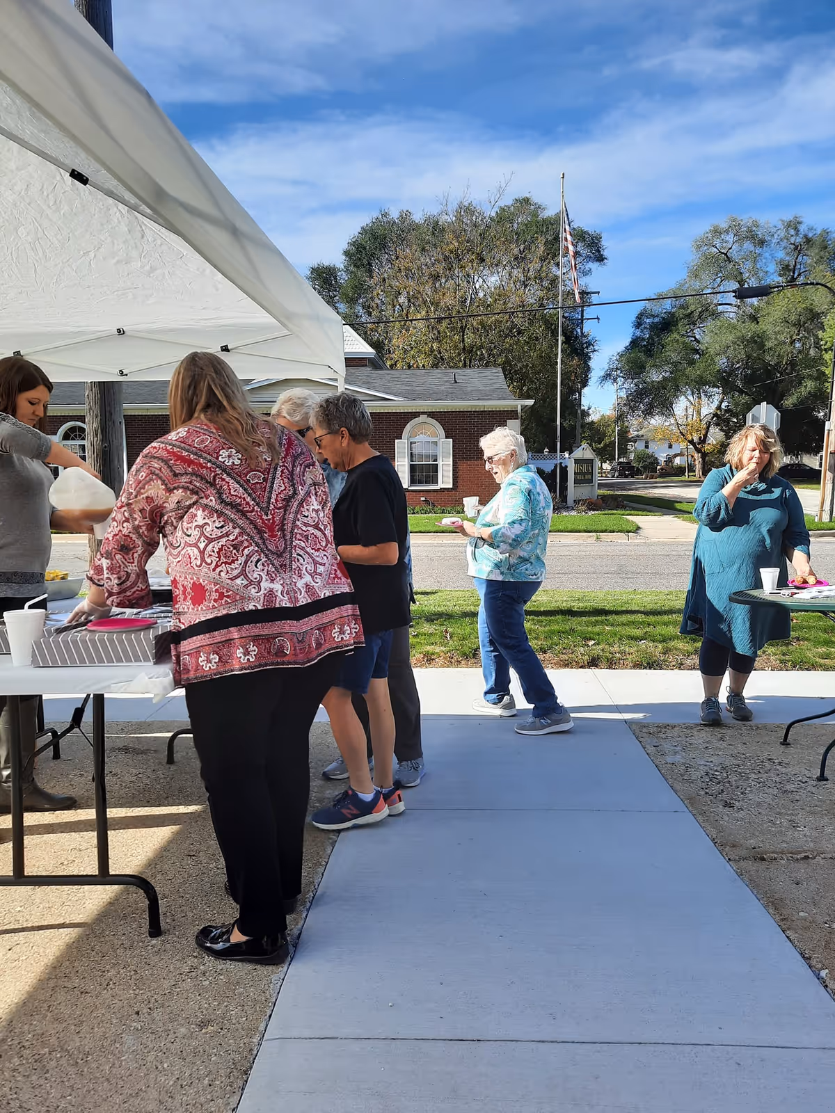 A group of elderly women gathered outside under a white canopy tent, serving and eating food on a sunny day. They are standing on a sidewalk with a brick building and trees in the background.