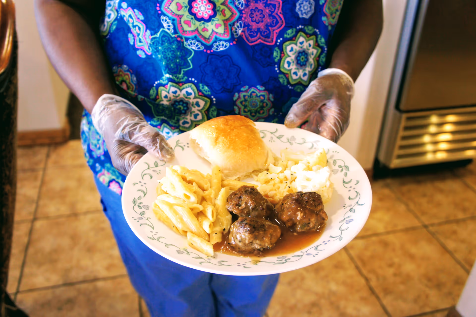 A person wearing a blue patterned shirt and disposable gloves is holding a plate of food that includes a bread roll, macaroni and cheese, three meatballs with gravy, and mashed potatoes.