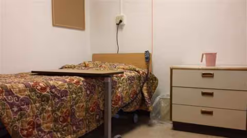 A sparsely furnished patient room with a patterned bed, dresser, and small table.