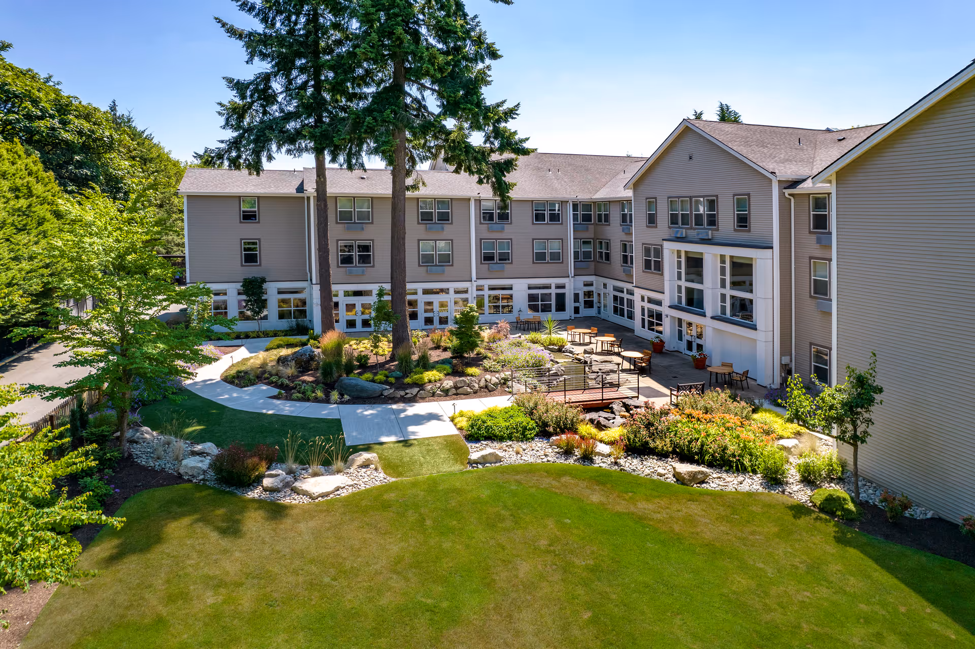 Outdoor view of GenCare Lifestyle Federal Way facility showing a landscaped garden with green grass, shrubs, and trees. The building surrounds the garden area with multiple windows and a patio with tables and chairs under a clear blue sky.