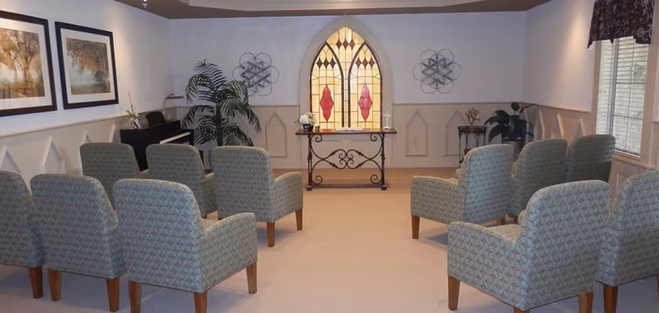 A small chapel or meditation room with two rows of upholstered chairs facing a decorative table with floral arrangements in front of a stained glass window. The room has framed artwork on the walls, a piano in the corner, and plants near the window and piano.