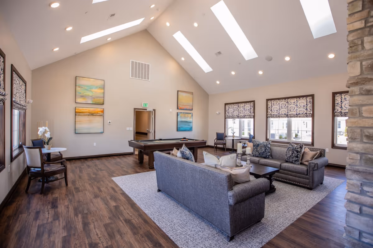 A spacious senior living common area with high vaulted ceilings and skylights. The room features two gray sofas with decorative pillows arranged around a dark wooden coffee table on a light gray rug. There is a pool table in the background, several windows with patterned Roman shades, and framed abstract paintings on the walls. The flooring is dark wood, and there is a stone pillar visible on the right side.