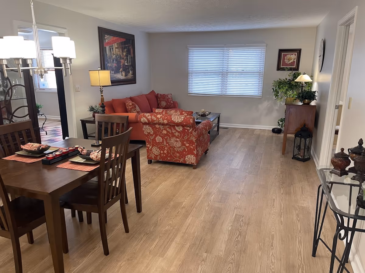 Open-plan living and dining area with wooden floors, a dark wood dining table, and red patterned sofa set beneath a window.
