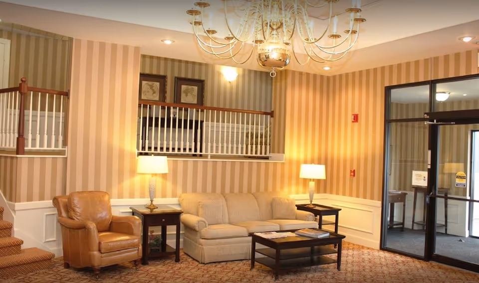 A warmly lit lobby-style seating area with a sofa, leather armchair, coffee table, side tables with lamps, and a chandelier against striped wallpaper.
