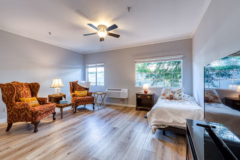 A bright senior living room with two patterned armchairs with pillows, a small wooden table with a lamp between them, a single bed with floral bedding and a teddy bear, a nightstand with a lamp, a large window showing greenery outside, a ceiling fan with light, and a flat-screen TV on a stand.