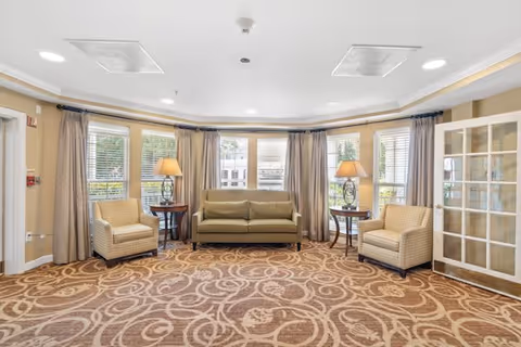 A bright and spacious living room area with a large patterned carpet, a green sofa centered between two beige armchairs, two side tables with lamps, and large windows with beige curtains letting in natural light.
