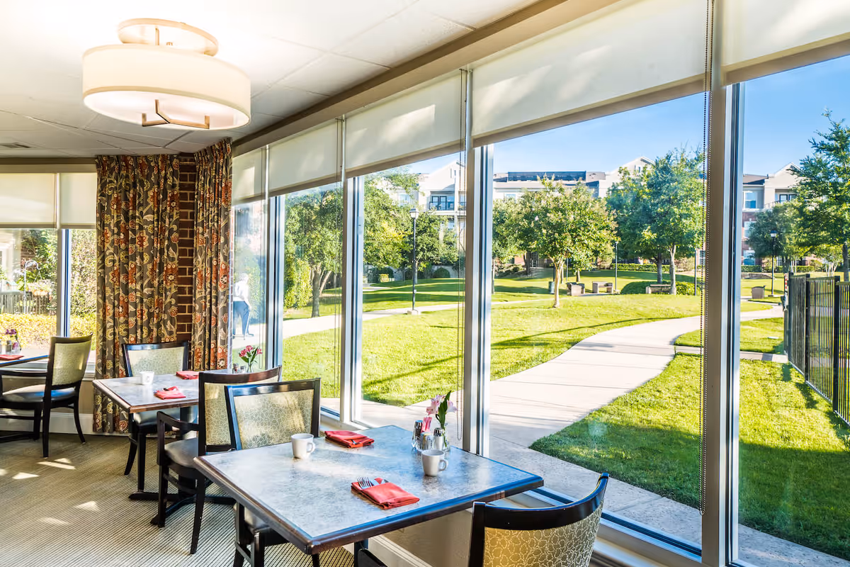Dining area with tables and chairs set with red napkins and white mugs, large windows providing a view of a green outdoor garden with trees and a walking path, and patterned curtains on one side.