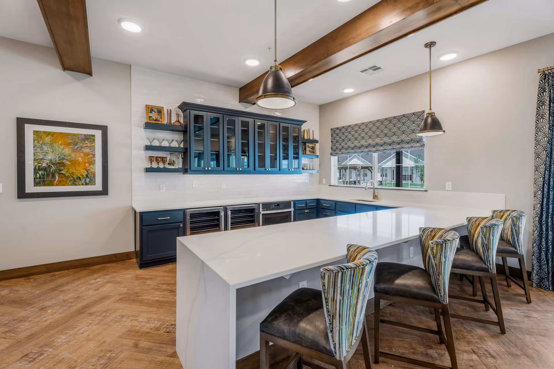 Modern kitchen area with a large white marble island countertop surrounded by four high-back chairs with patterned upholstery. The kitchen features dark blue cabinets with glass doors, open shelves displaying glassware, and two built-in wine coolers. A window with patterned blinds overlooks an outdoor area. The room has wooden flooring, exposed wooden ceiling beams, and two pendant lights hanging above the island.